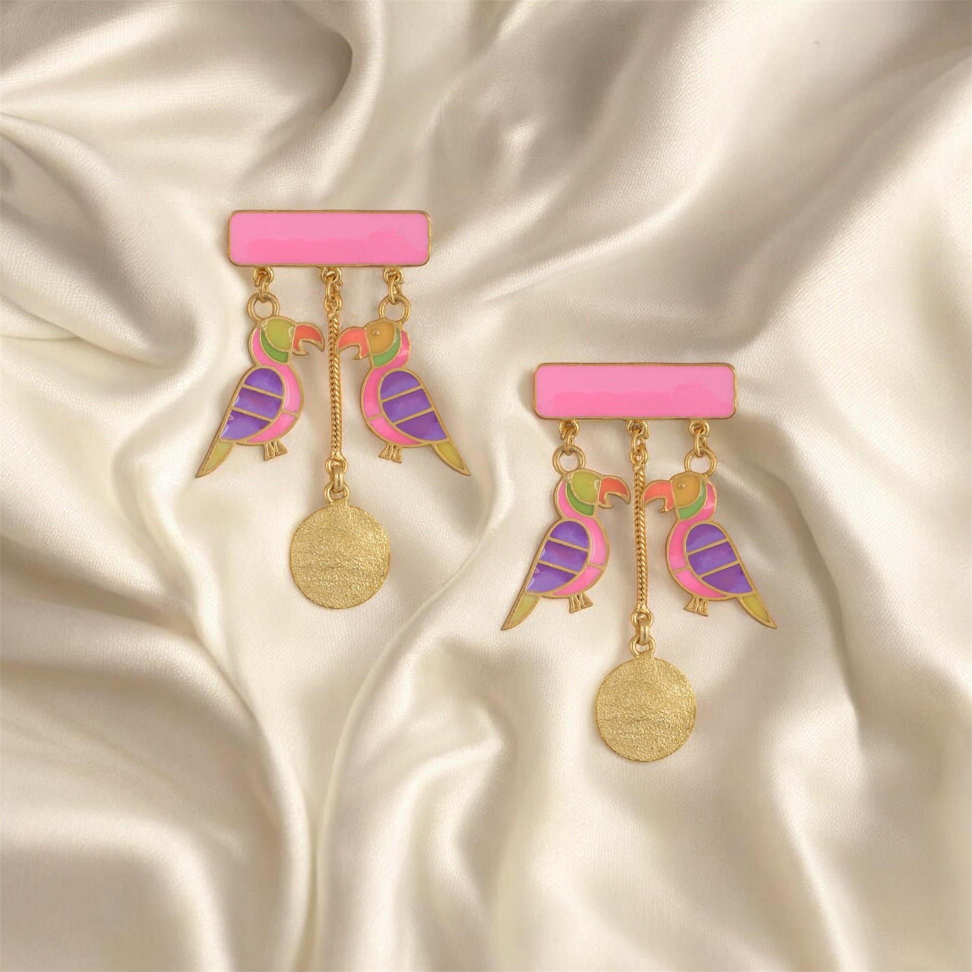 Colorful bird-shaped earrings on a pink bar with gold accents, set against a satin background.