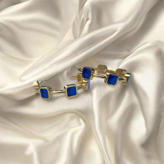 Gold hoop earrings with blue square stones on a satin surface